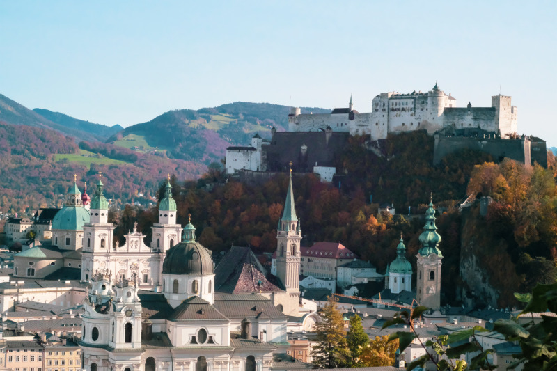 Blick auf die historische Festung Hohensalzburg auf einem Hügel über der Stadt, mit der barocken Architektur Salzburgs im Vordergrund und bewaldeten Hügeln im Hintergrund bei Tageslicht.