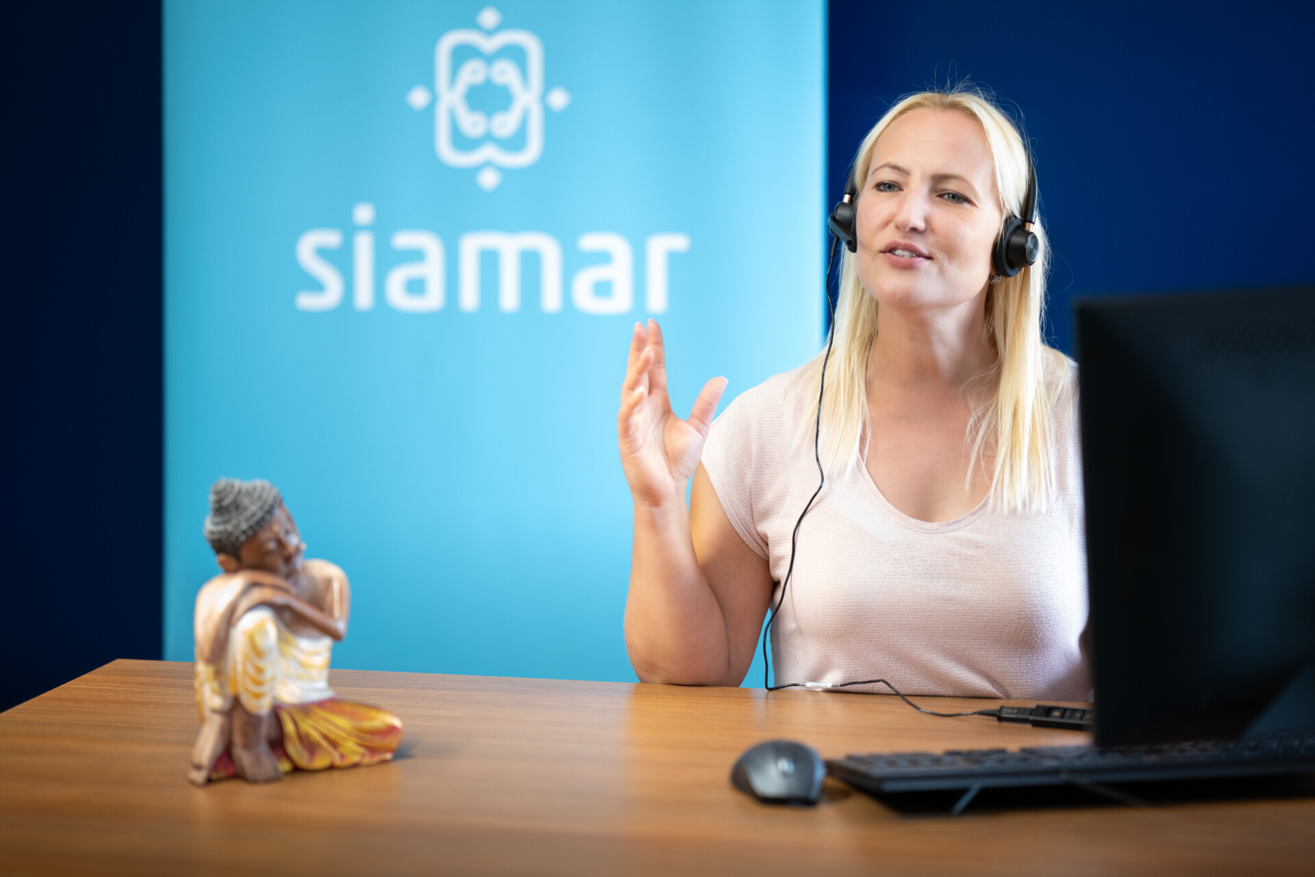 Eine blonde Frau mit Headset sitzt an einem Schreibtisch und gestikuliert während eines Gesprächs, vermutlich in einem Call-Center. Vor ihr steht eine kleine, farbenfrohe Figur. Im Hintergrund ist ein Banner mit dem Logo "siamar" zu sehen.
