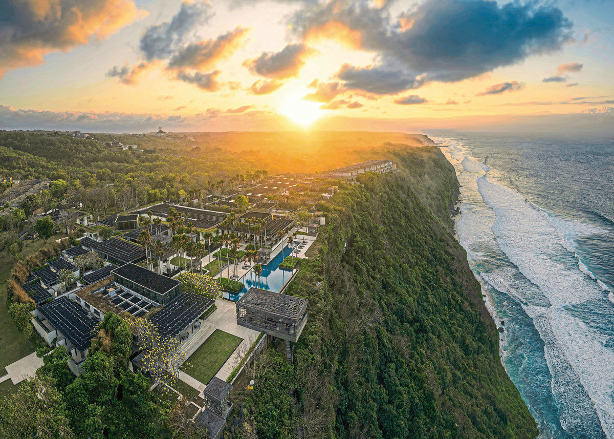 Luftaufnahme eines luxuriösen Resortkomplexes am Rande einer hohen, bewachsenen Klippe bei Sonnenuntergang; der Blick erstreckt sich über reihenweise Villendächer zum schimmernden Meer, das mit weißen Wellen gegen die Küste schlägt.