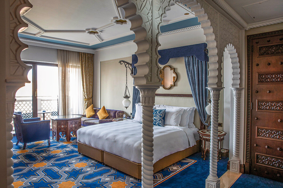 Luxuriöses Schlafzimmer im orientalischen Stil mit kunstvoll gearbeitetem Baldachinbett, ornamentierten Säulen und leuchtend blauem Teppich mit geometrischen Mustern.