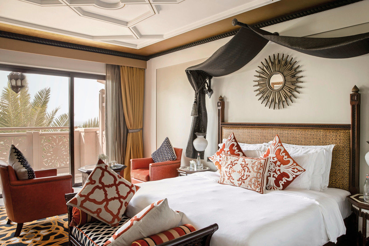 Ein luxuriös eingerichtetes Hotelzimmer im marokkanischen Stil mit einem großen Doppelbett, bestückt mit zahlreichen Kissen mit ornamental gemusterten Bezügen in Rot- und Orangetönen. Über dem Bett ist ein schwarzes Stoffsegel angebracht, das an zwei kunstvoll geschnitzten Halterungen hängt. An der Wand prangt ein dekorativer Spiegel mit einem strahlenförmigen Rahmen. Vor dem Fenster, durch das eine Palme und ein blauer Himmel sichtbar sind, stehen zwei bequeme, rote Sessel und ein kleiner Beistelltisch. Der Raum zeichnet sich durch eine Mischung aus traditionellen Elementen und modernem Komfort aus.