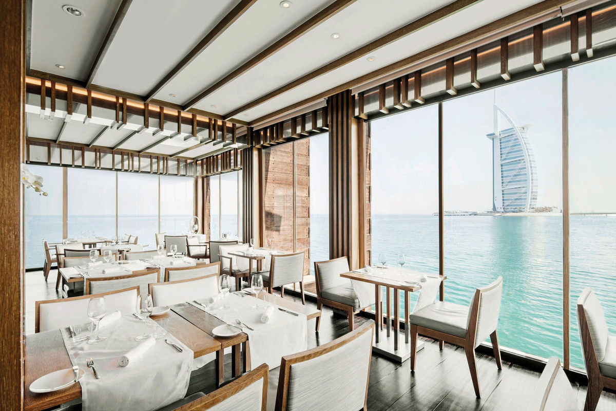 Ein elegantes Restaurant mit moderner Einrichtung und bodentiefen Fenstern, die einen beeindruckenden Blick auf das Meer und ein ikonisches, segelförmiges Gebäude im Hintergrund bieten. Die Tische sind mit weißen Tischdecken, Besteck und Gläsern für ein gehobenes Esserlebnis liebevoll eingedeckt.