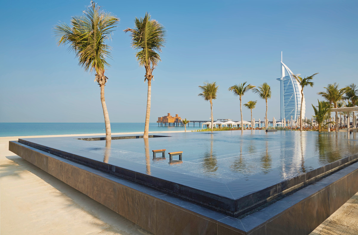 Ein luxuriöses Infinity-Pool mit klarem Wasser, umgeben von Palmen, mit Blick auf einen ruhigen, blauen Ozean und ein ikonisches, segelförmiges Gebäude im Hintergrund bei klarem Himmel.