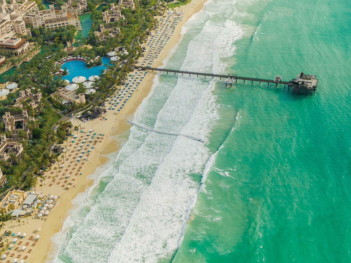 Luftaufnahme eines tropischen Strandresorts mit einem Pier, der sich ins Meer erstreckt, umgeben von türkisfarbenem Wasser und Wellen, die sanft auf den sandigen Strand treffen.