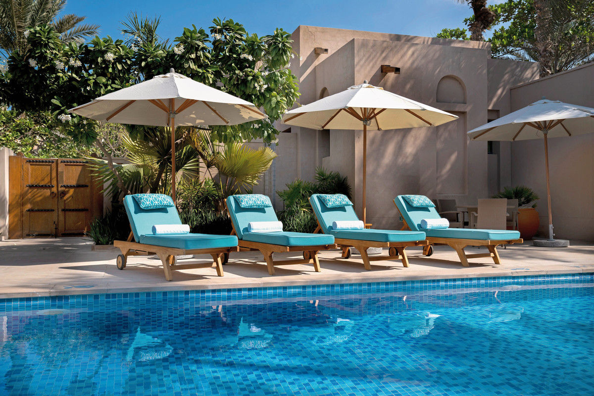 Ein idyllischer Poolbereich mit klarem blauem Wasser vor einem traditionellen Gebäude, umgeben von üppiger Vegetation. Vier türkisfarbene Liegestühle mit passenden Handtüchern sind unter zwei weißen Sonnenschirmen aufgestellt. Die Szene strahlt Ruhe und Entspannung aus.