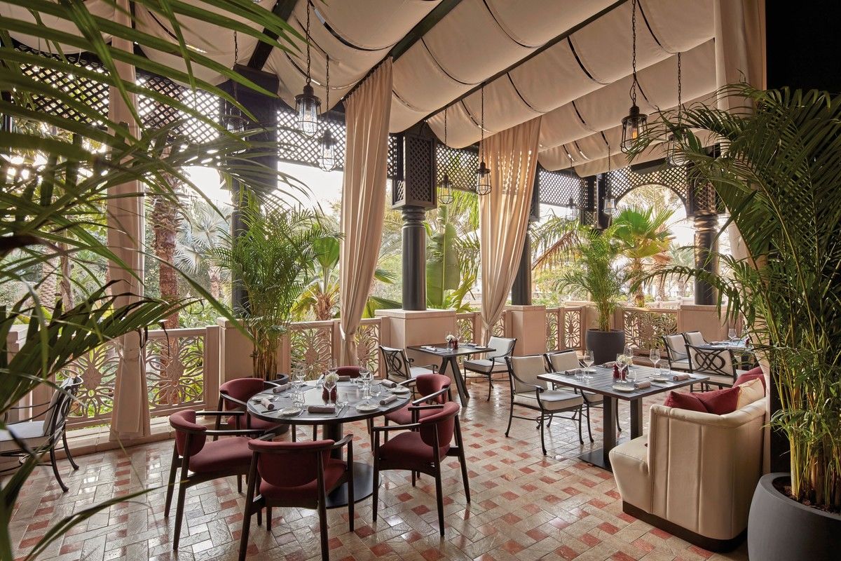Elegantes Restaurant mit offenem Ambiente, umgeben von üppigem Grün und Palmen, mit einem aufwendigen Mosaikboden, stilvollen Tischen und Stühlen in Rot- und Beigetönen sowie sorgfältig arrangiertem Geschirr. Sonnenlicht scheint durch Deckenfenster und schafft eine warme und einladende Atmosphäre.