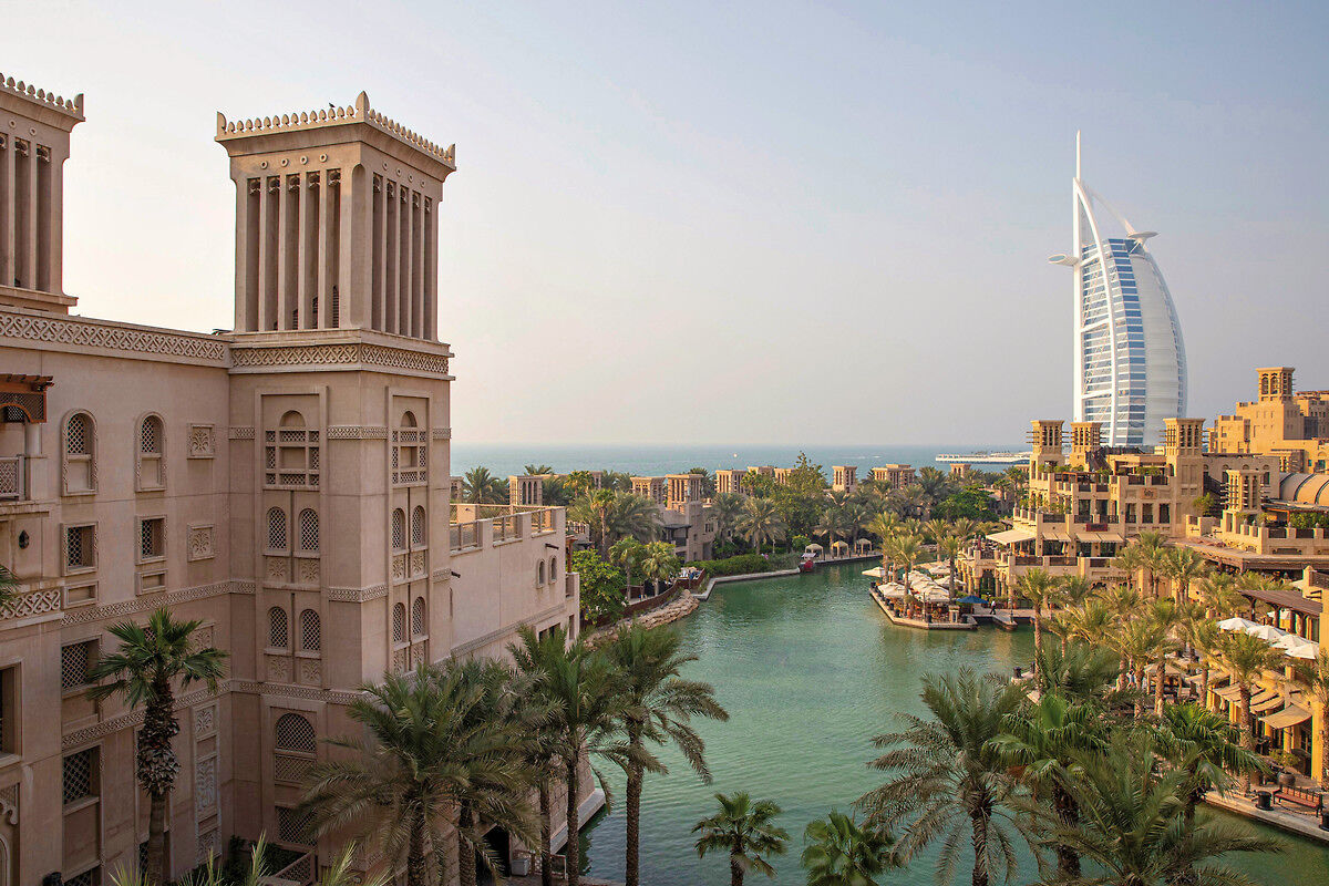 Blick auf eine idyllische Wasserstraße in Dubai, umgeben von Palmen und traditioneller Architektur im Vordergrund und dem ikonischen Burj Al Arab Hotel im Hintergrund bei Tageslicht.