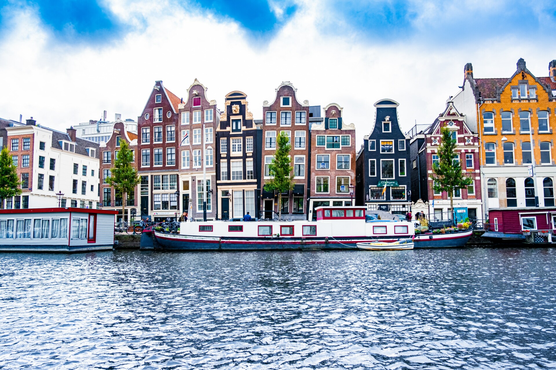 Blick auf traditionelle niederländische Häuser mit Giebelfassaden in einer Reihe an einem Kanal in Amsterdam. Im Vordergrund ist das schimmernde Wasser des Kanals, vor den Häusern liegen Hausboote vertäut. Wolken und ein blauer Himmel vervollständigen die Szenerie.