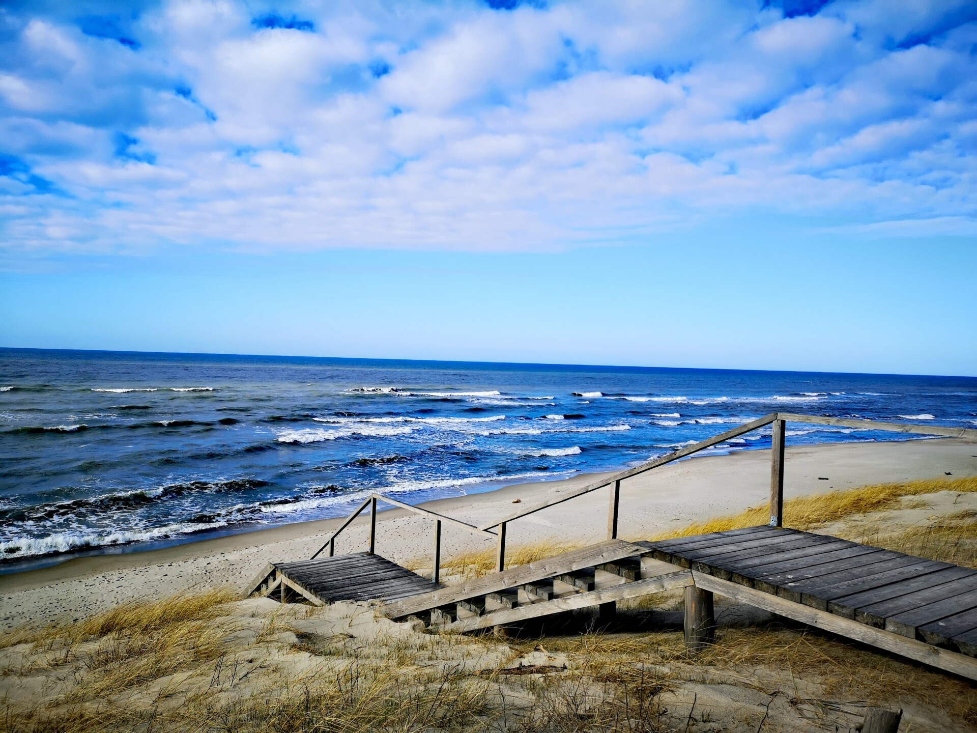 Bild eines ruhigen Strandes mit einer Holztreppe, die durch Dünen zum Meer führt, unter einem blauen Himmel mit leichter Bewölkung.