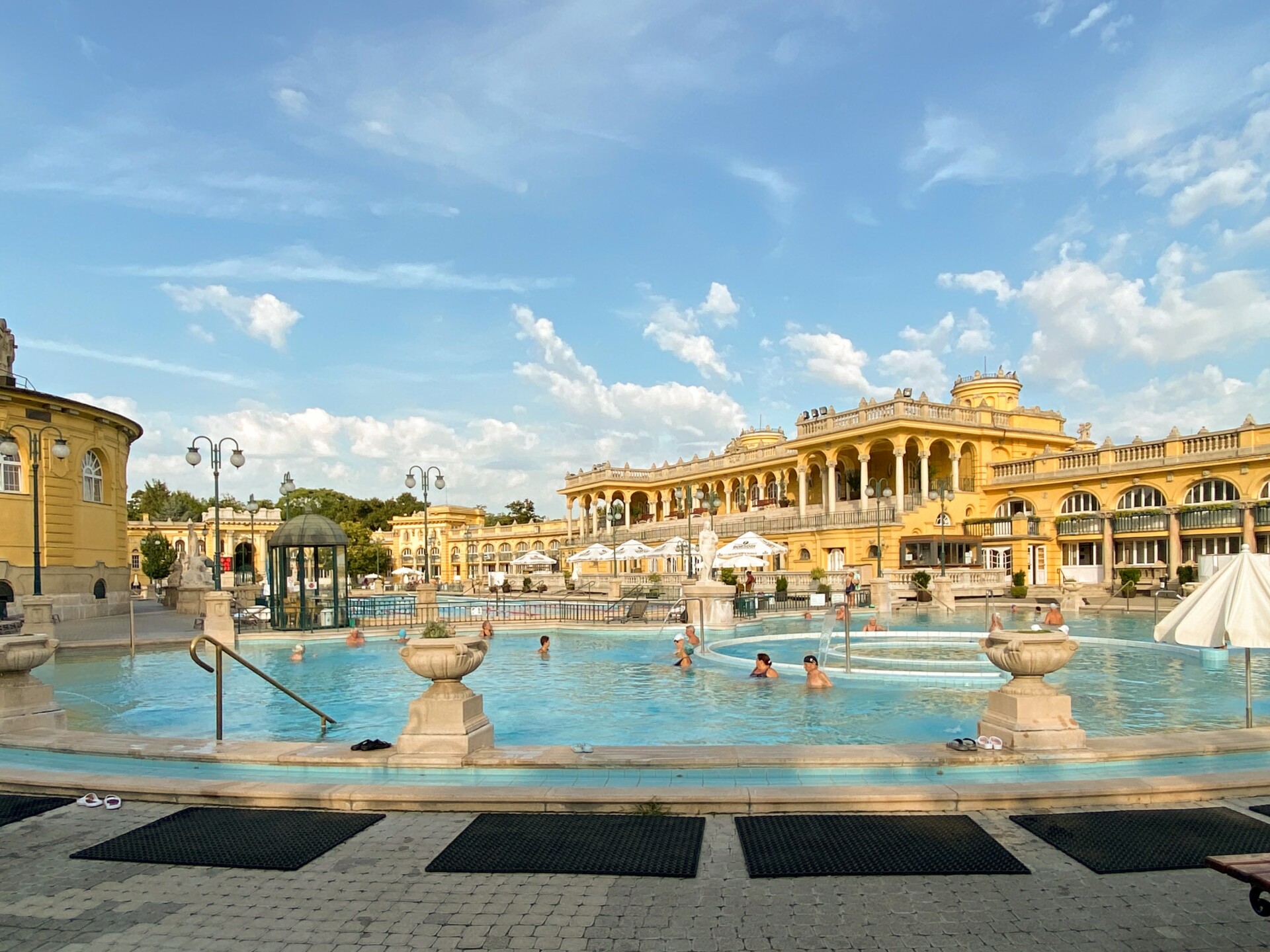Ein weitläufiges Freibad mit klarem blauem Wasser, umgeben von neoklassizistischen gelben Gebäuden mit Bogengängen und Verzierungen. Mehrere Personen entspannen sich im Wasser und auf den Poolrändern an einem sonnigen Tag mit vereinzelten Wolken am Himmel.