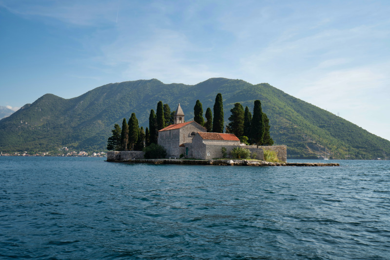 Eine kleine Insel mit einer historischen Kirche und Zypressen, umgeben vom blauen Wasser eines Sees, vor der Kulisse grüner Berge unter einem klaren, blauen Himmel.