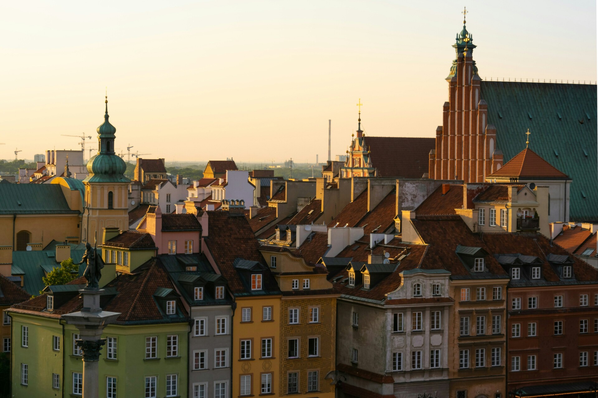 Blick auf die Dächer einer europäischen Altstadt bei Sonnenuntergang, mit historischen Gebäuden in verschiedenen warmen Farbtönen und einigen Kirchtürmen, die sich gegen den orange gefärbten Himmel abzeichnen.