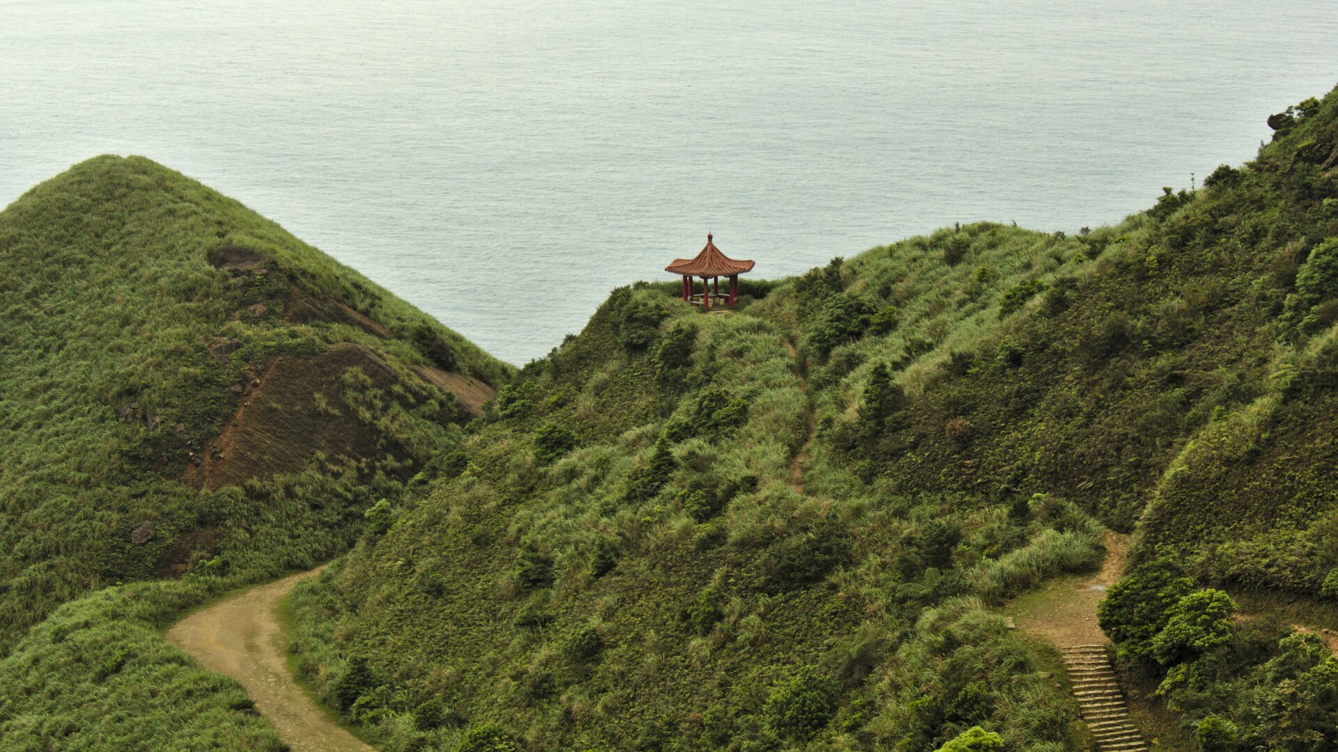 Ein traditioneller Pavillon sitzt auf einem grünen, grasbewachsenen Hügel über dem Meer. Ein gewundener Pfad führt zum Pavillon hoch, umgeben von üppigem Grün und der ruhigen Wasserfläche im Hintergrund.