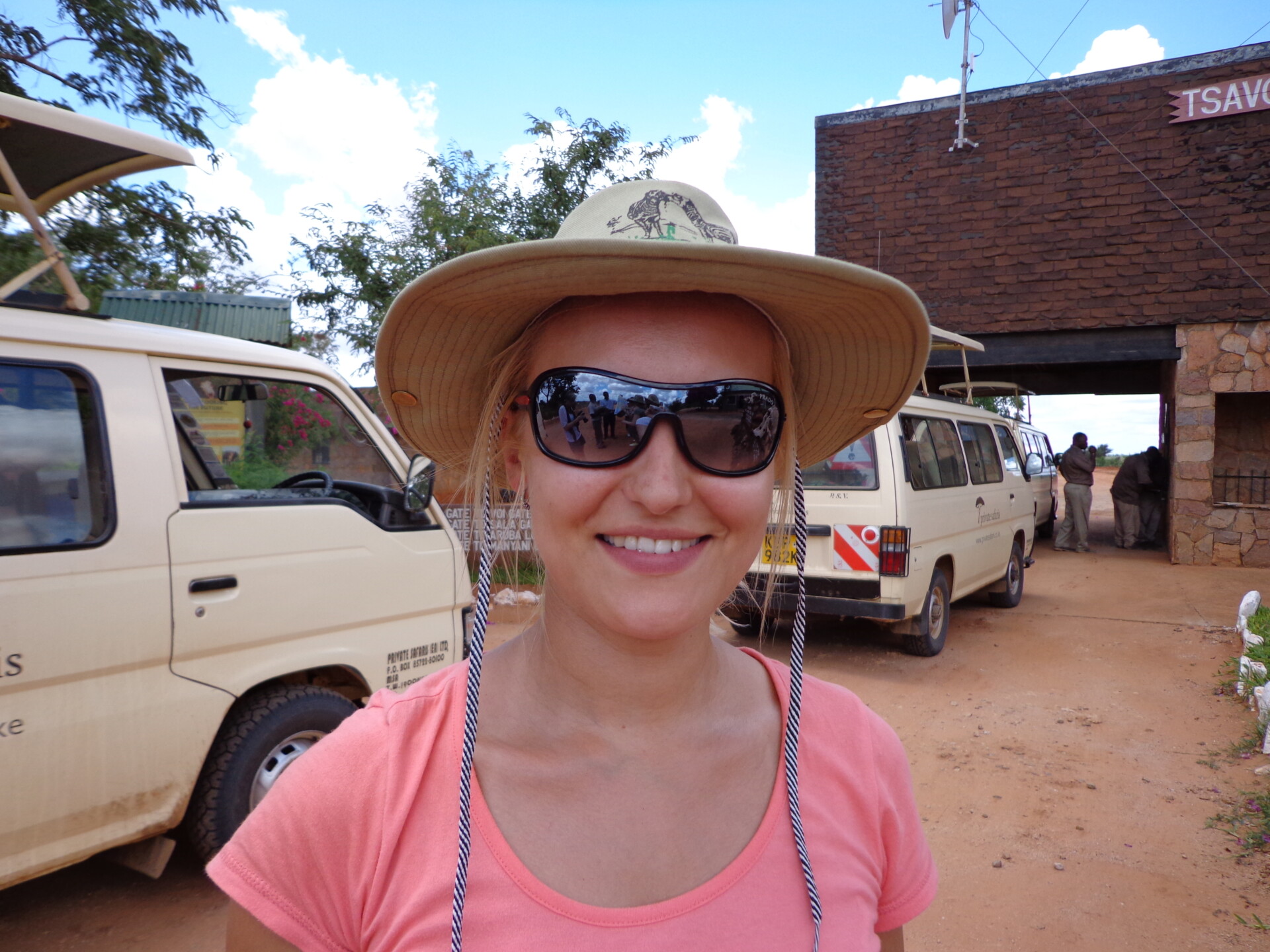 Eine lächelnde Frau mit Sonnenbrille und breitkrempigem Hut steht vor einem Hintergrund mit Safarifahrzeugen und einem Gebäude mit der Aufschrift "TSAVO". Sie scheint sich auf ein Abenteuer in einem Naturreservat oder einer ähnlichen Umgebung vorzubereiten.