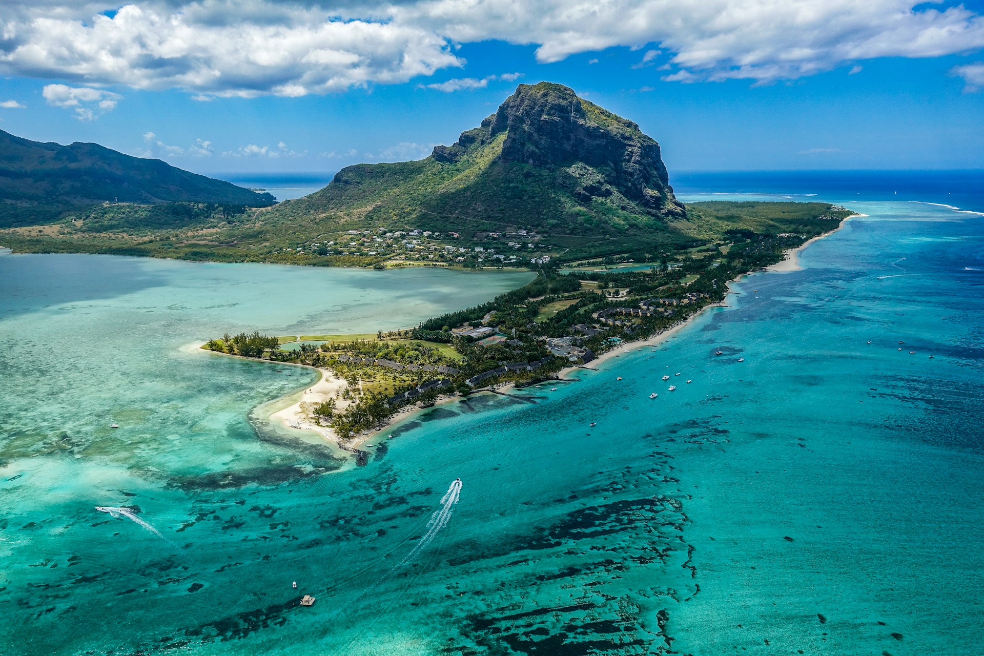 Luftaufnahme eines beeindruckenden tropischen Bergs neben einer Küstenlinie mit klarem türkisfarbenem Wasser, Korallenriffen und Booten, die nahe einem Strand mit Palmen und Unterkünften zu sehen sind.