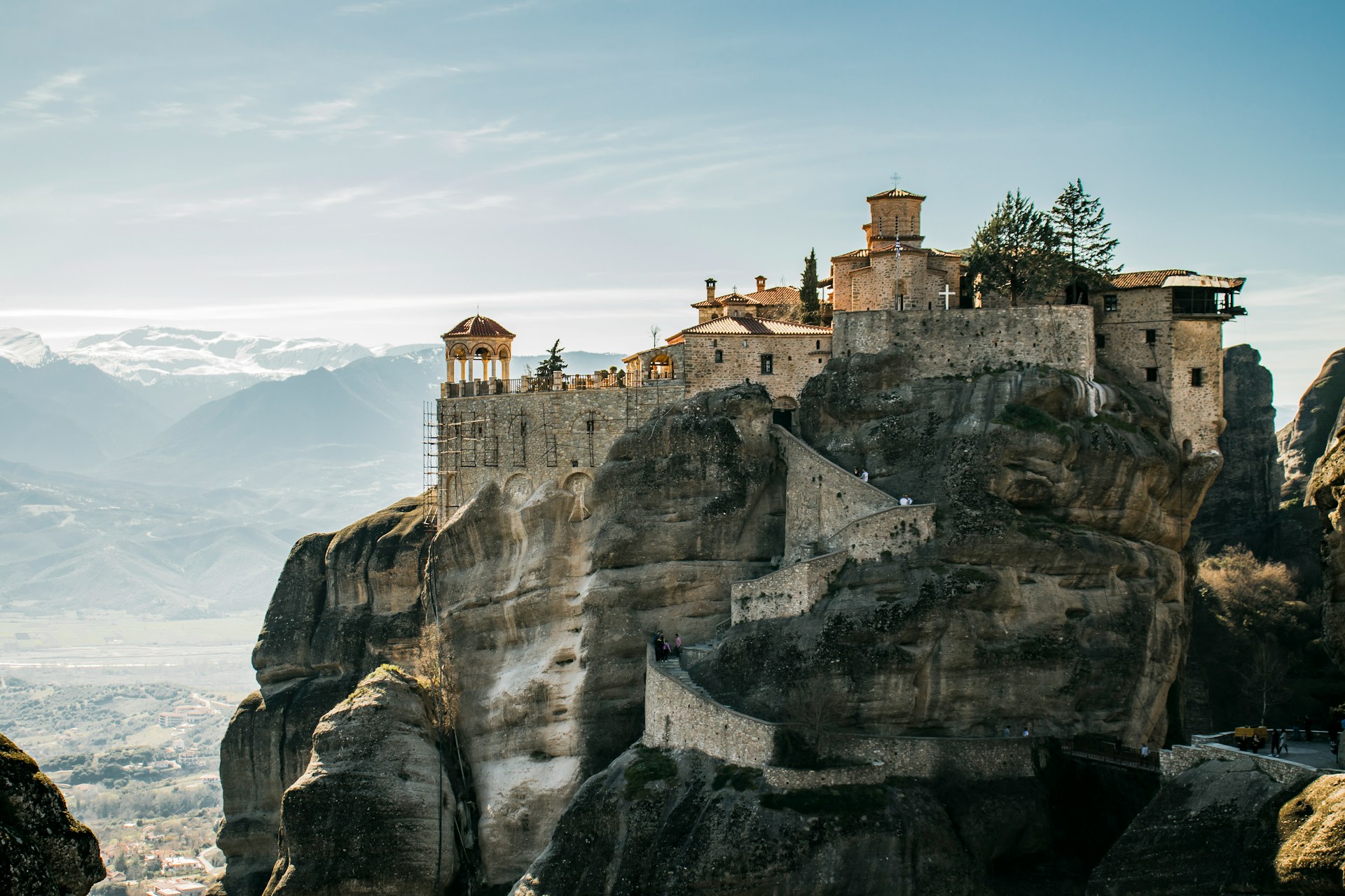 Alt-Text: Eine historische Klosteranlage, erbaut auf einem hohen Felsvorsprung, mit einem beeindruckenden Panoramablick auf umliegende Bergketten und das darunter liegende Tal im Sonnenlicht.
