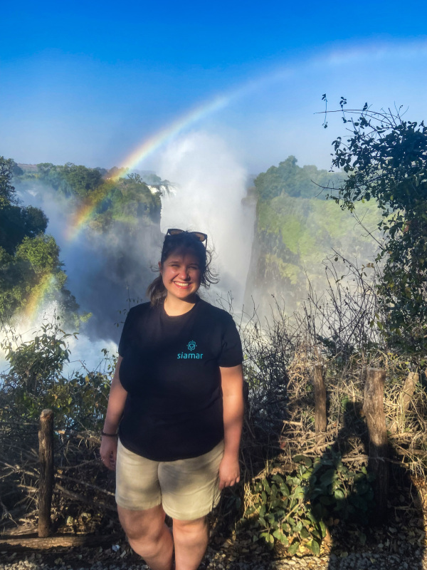 Eine lächelnde Person steht im Vordergrund und trägt eine Sonnenbrille auf dem Kopf sowie ein schwarzes T-Shirt mit einem Logo. Im Hintergrund sieht man eindrucksvolle Wassermassen, die von einem Wasserfall herabstürzen und einen Regenbogen erzeugen, der sich vor einer bewaldeten Klippe spannt.