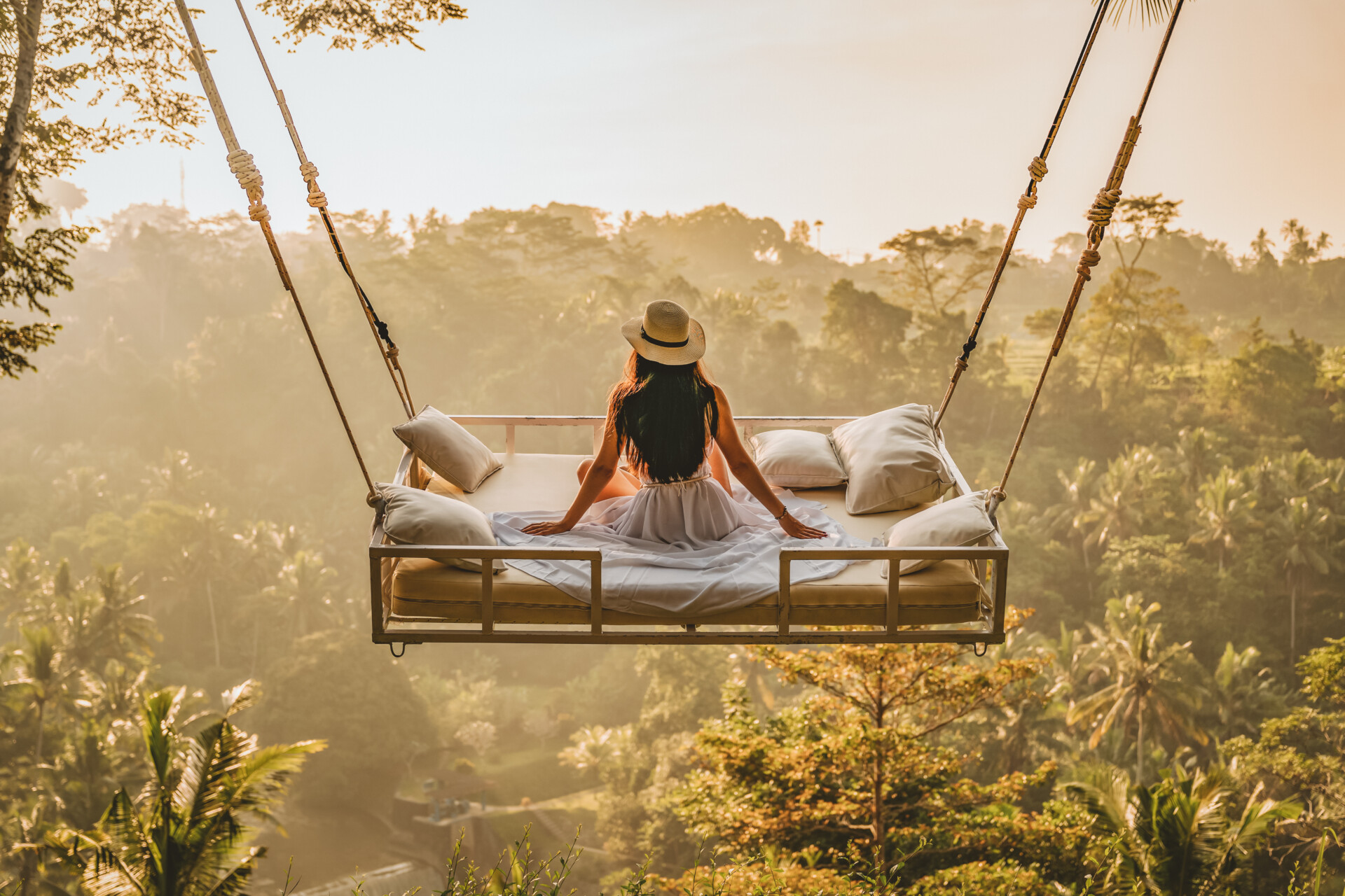 Eine Person sitzt mit dem Rücken zur Kamera auf einem großen Hängebett im Freien, umgeben von einem tropischen Wald. Die Morgensonne taucht die Szene in ein sanftes Licht. Die Person trägt einen Strohhut und blickt auf die üppigen Bäume und die Landschaft im Hintergrund.