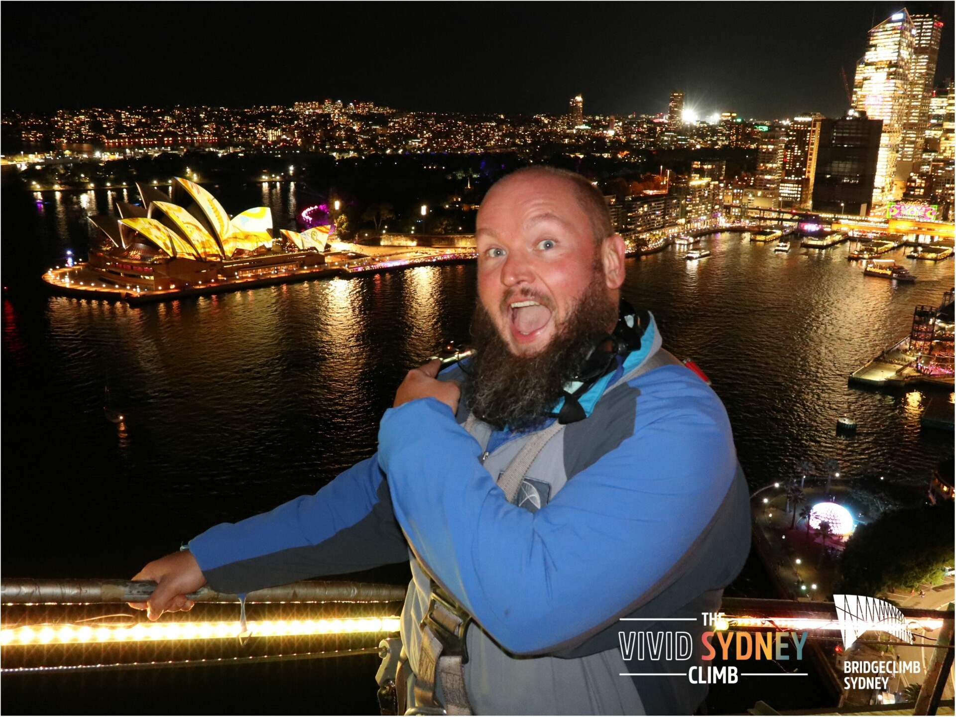 Alternativtext: Mann mit Bart in blauer und grauer Kletterausrüstung posiert glücklich vor der nächtlichen Skyline von Sydney mit betonter Beleuchtung des Sydney Opera House, dem Hafen und umliegenden Gebäuden. Werbung für "The Vivid Sydney Climb" und "BridgeClimb Sydney" ist ebenfalls sichtbar.