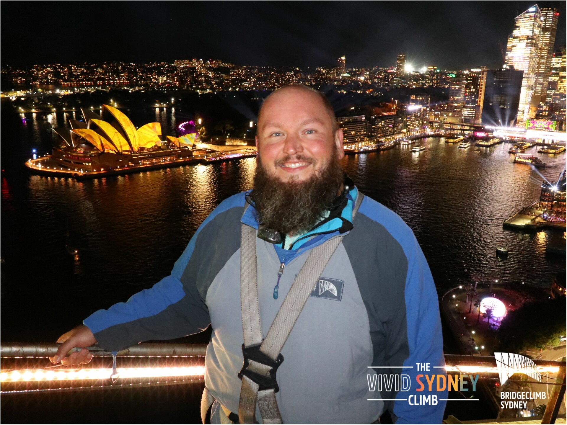 Ein lächelnder Mann mit Bart in Sicherheitsausrüstung steht auf einer Aussichtsplattform bei Nacht. Im Hintergrund ist die beleuchtete Skyline von Sydney mit dem markanten Sydney Opera House und der Hafenbrücke zu sehen. Lichter reflektieren im Wasser des Hafens, und die Stadt wirkt lebhaft beleuchtet. Textelemente im Bild weisen auf "The Vivid Sydney Climb" und "Bridgeclimb Sydney" hin.