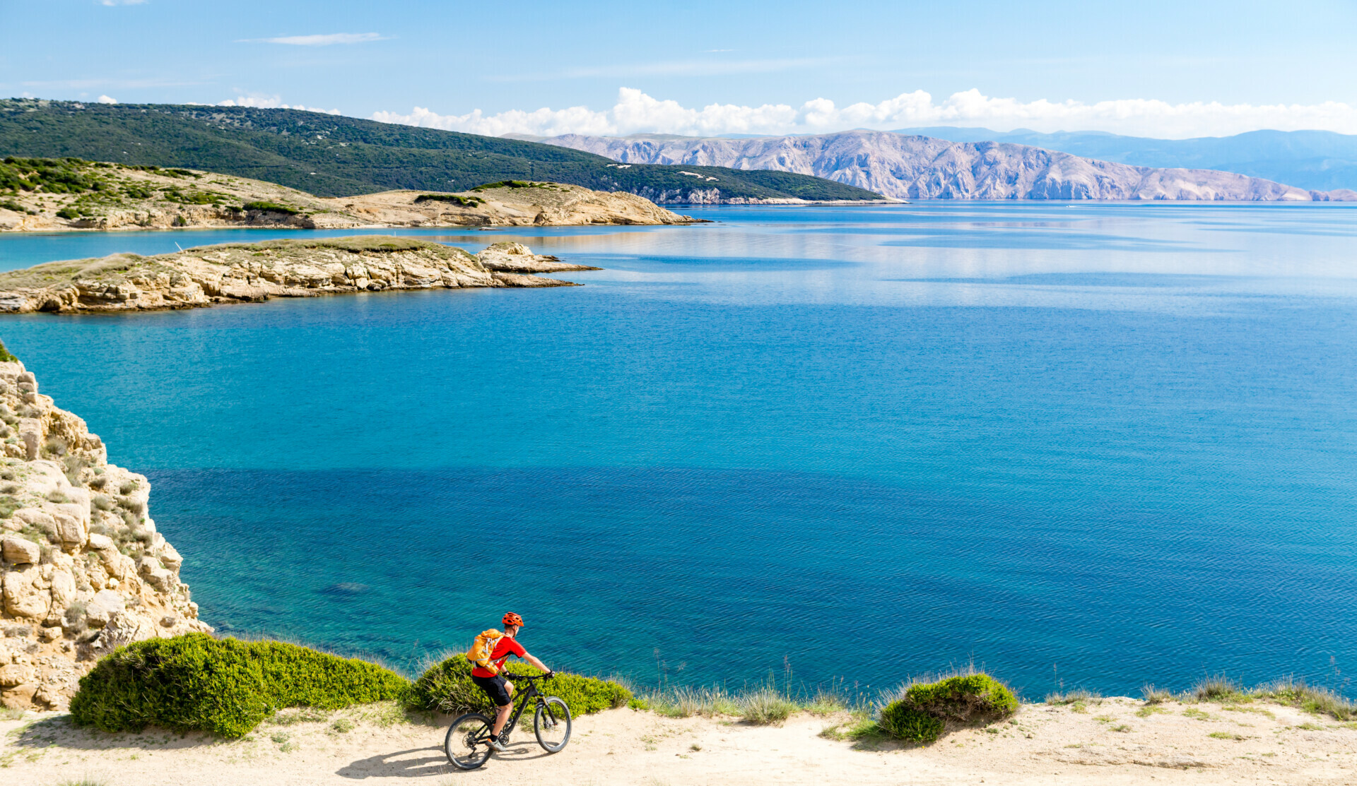 Ein Radfahrer in leuchtender Sportkleidung fährt auf einem sandigen Weg entlang der malerischen Küstenlinie, mit klarem blauem Meerwasser und einer bergigen Landschaft im Hintergrund.