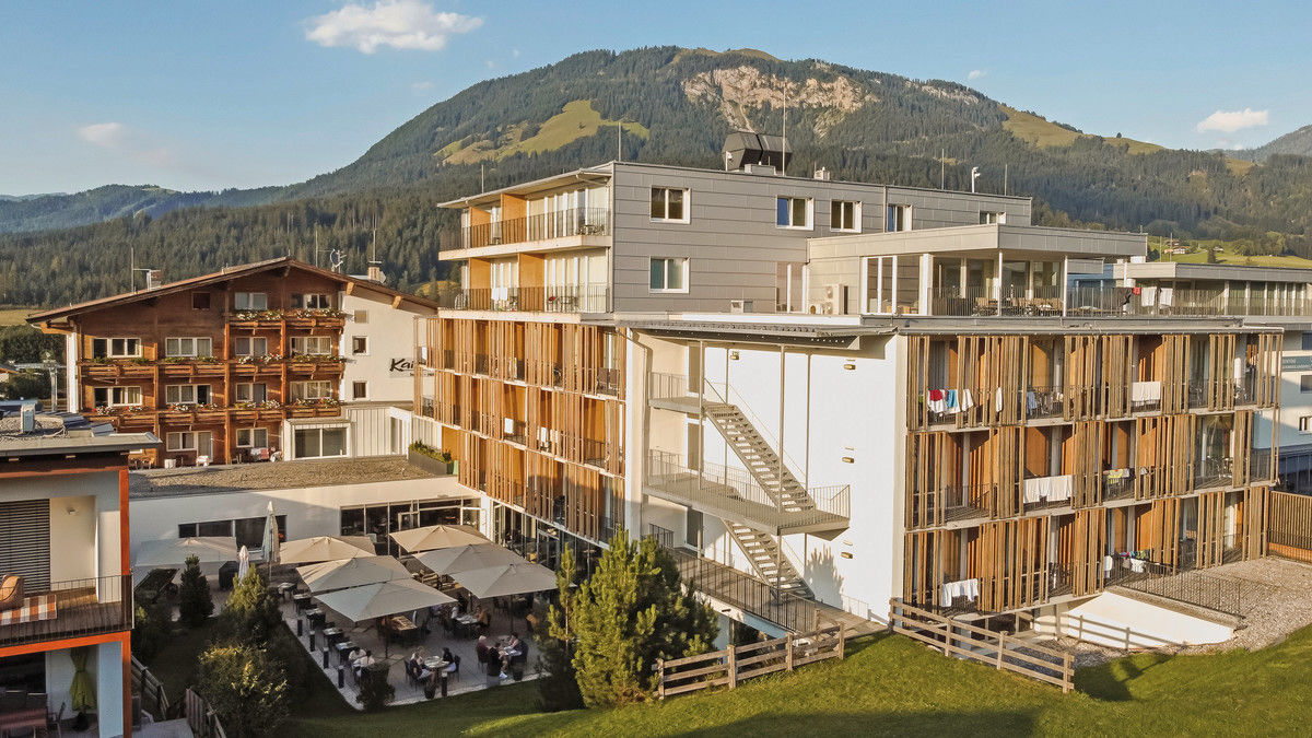 Ein modernes Hotelgebäude mit Holzfassaden neben einem traditionellen, alpenländischen Gasthaus mit blumenbestückten Balkonen. Im Vordergrund befindet sich eine Terrasse mit Sitzgelegenheiten und Sonnenschirmen, dahinter erstreckt sich eine grüne Wiese. Im Hintergrund erheben sich bewaldete Berge unter einem blauen Himmel.