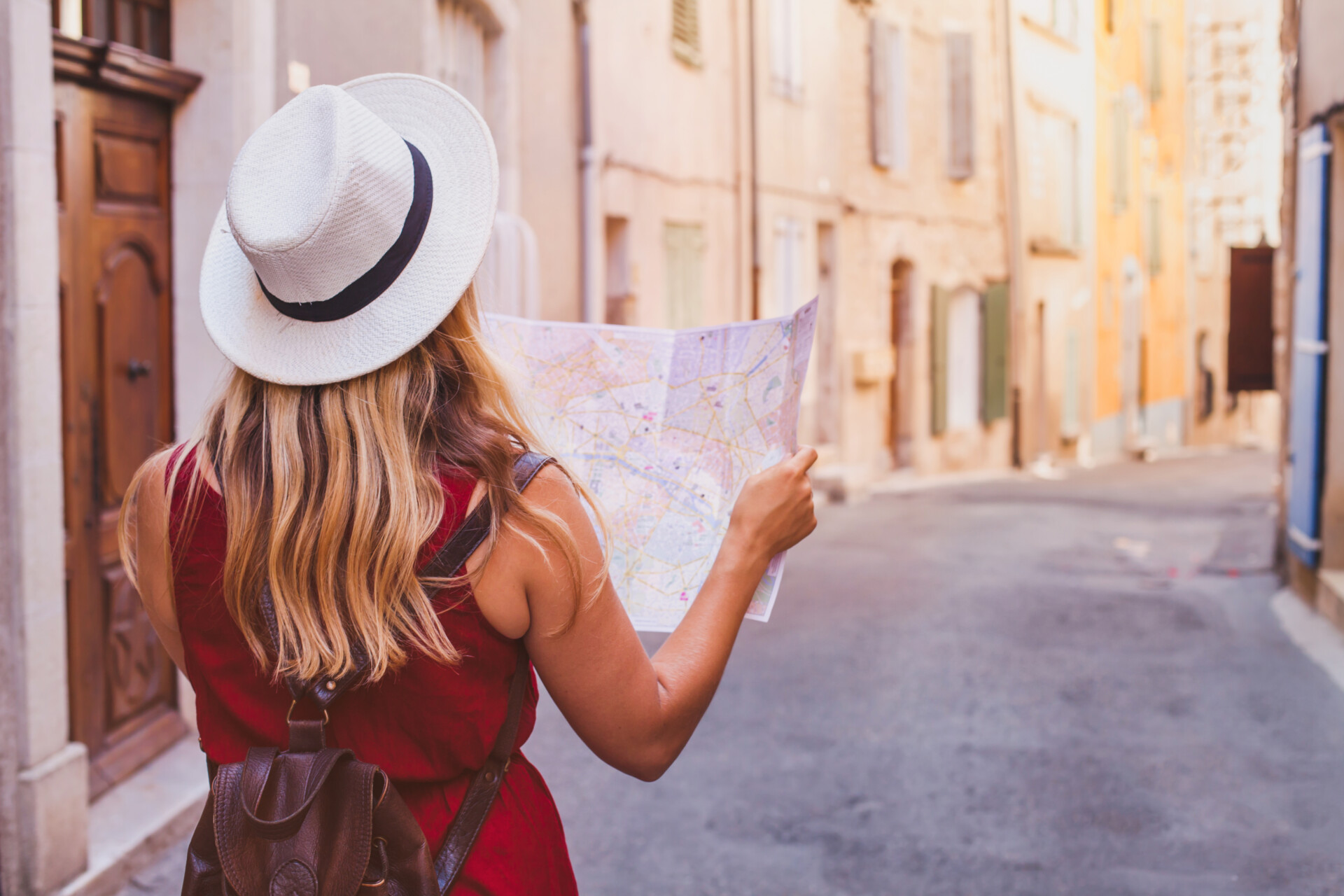 Eine Frau, von hinten betrachtet, die ein rotes Kleid trägt und einen weißen Hut mit schwarzer Krempe auf hat, steht in einer ruhigen Straße und hält eine geöffnete Landkarte. Sie scheint den Weg in einer Altstadt mit pastellfarbenen Gebäuden zu suchen.