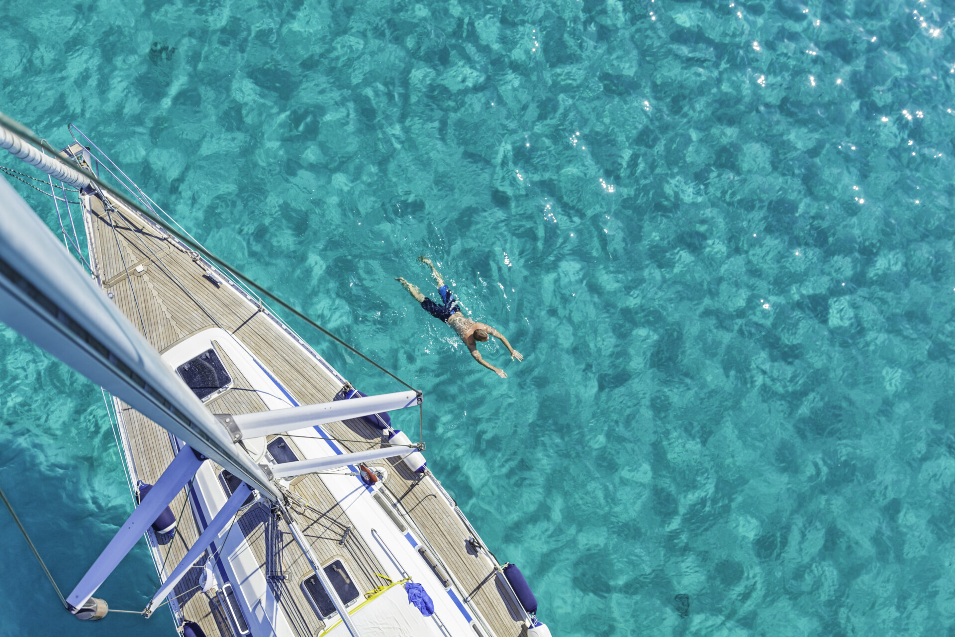 Eine Draufsicht auf eine Person, die neben einem Katamaran in klarem, türkisfarbenem Wasser schwimmt. Das Deck des weißen Segelschiffs mit blauen Akzenten ist sichtbar, während die Person entspannt im Wasser treibt.