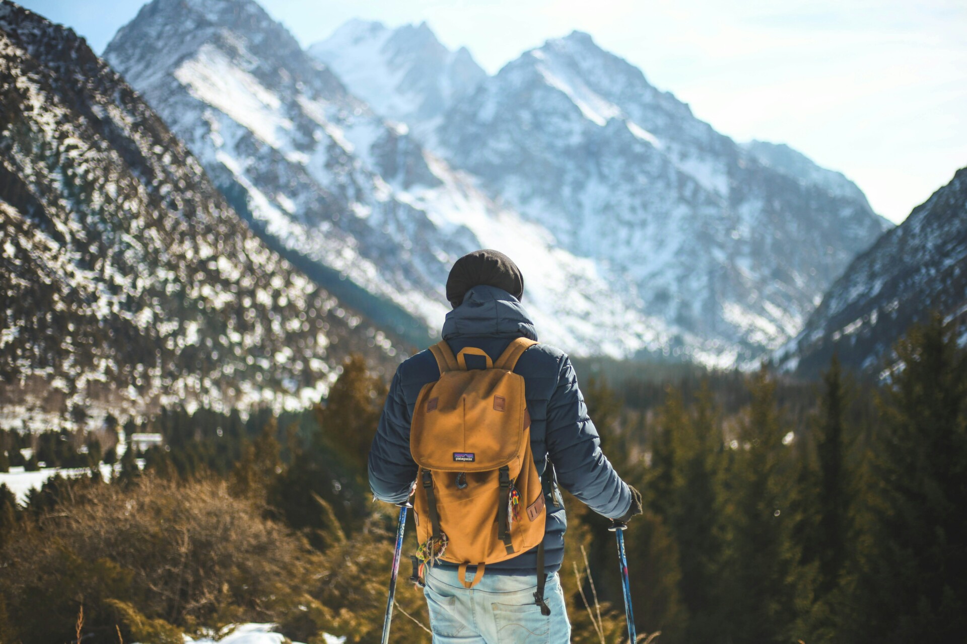 Eine Person mit Rucksack und Wanderstöcken steht vor einer beeindruckenden, schneebedeckten Bergkulisse und blickt in die Ferne.